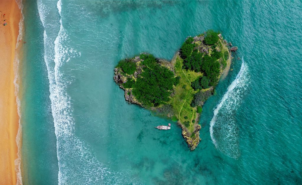 Heart-shaped tropical island surrounded by turquoise waters, with a sandy beach and a boat nearby.