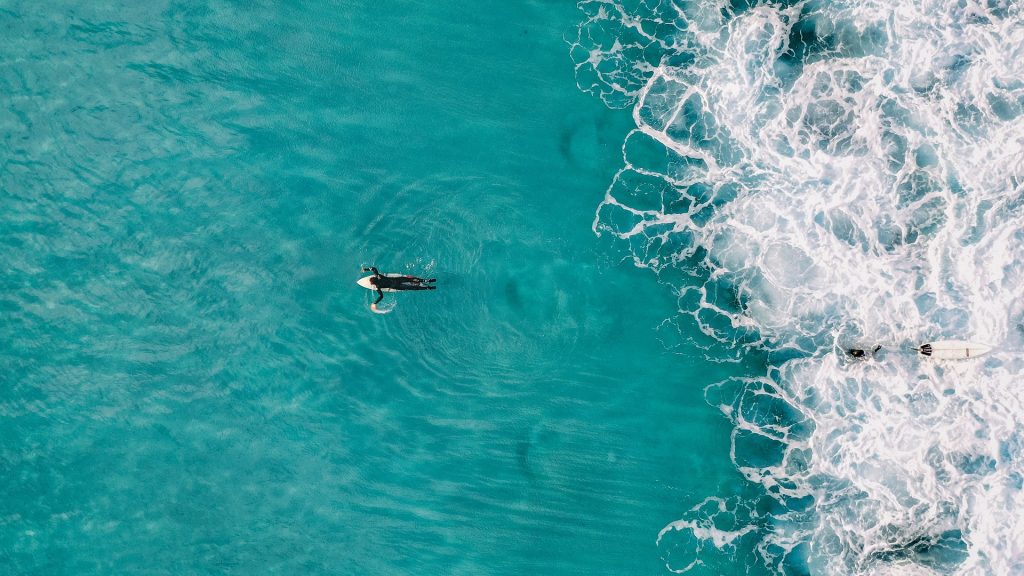 A Bird's eye view of a person paddling in clear blue sea, a wave following them close behind.