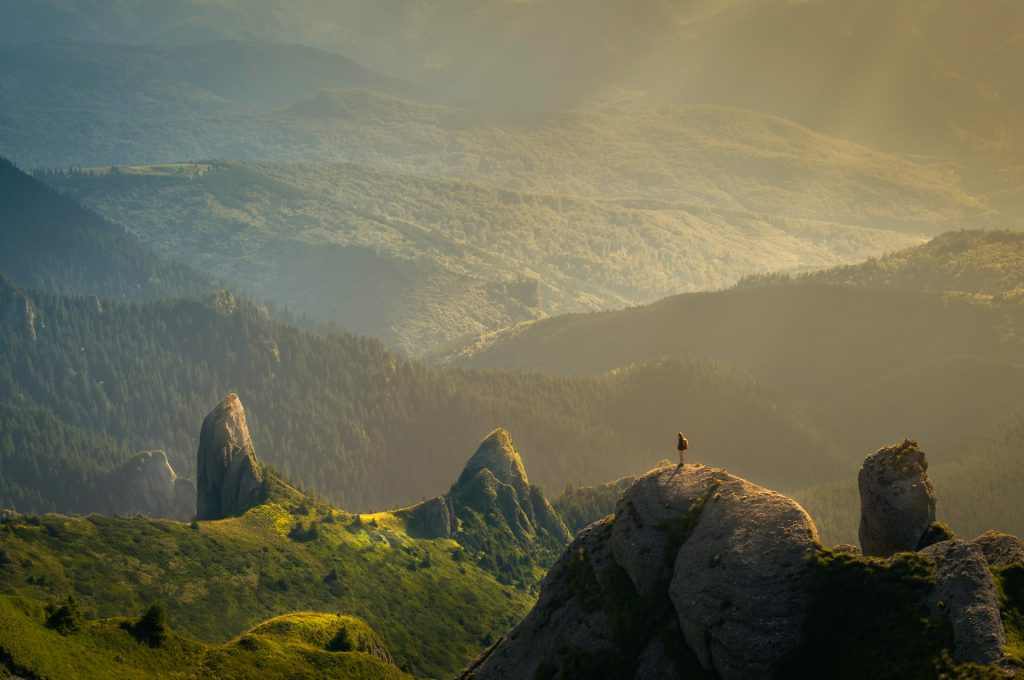 A hiker stands atop a summit, looking out over a verdant lanscape and a range of hills and mountains.