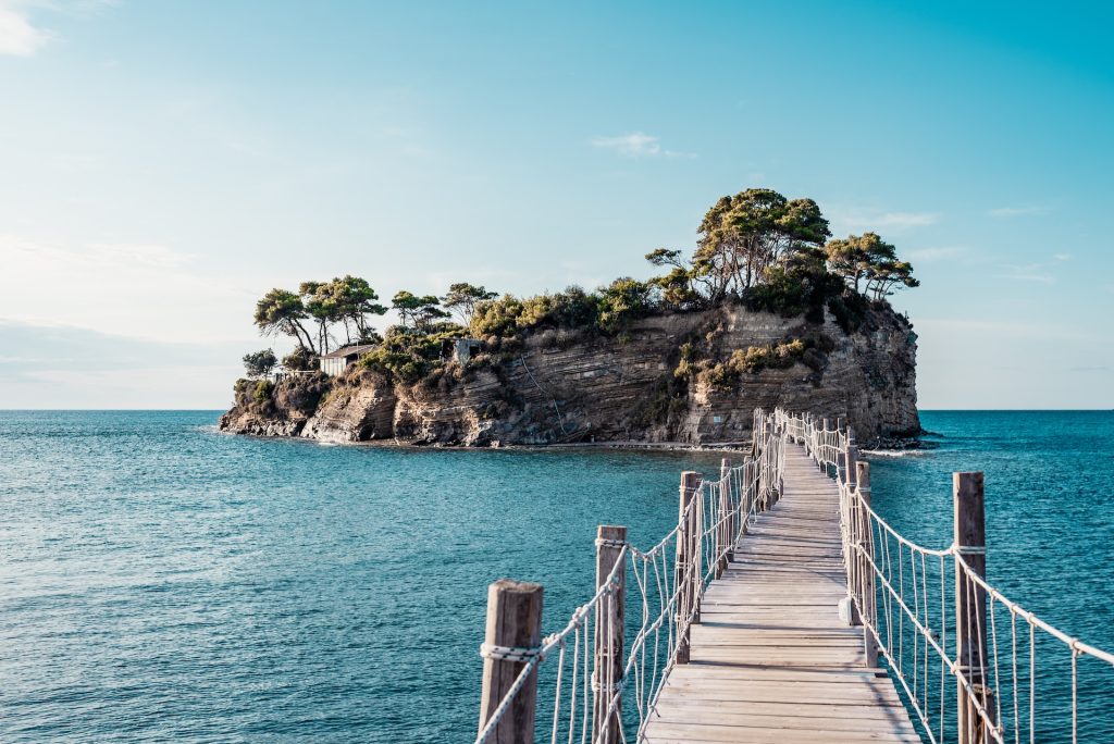 A first person view of a wooden bridge leading over blue water to an island.