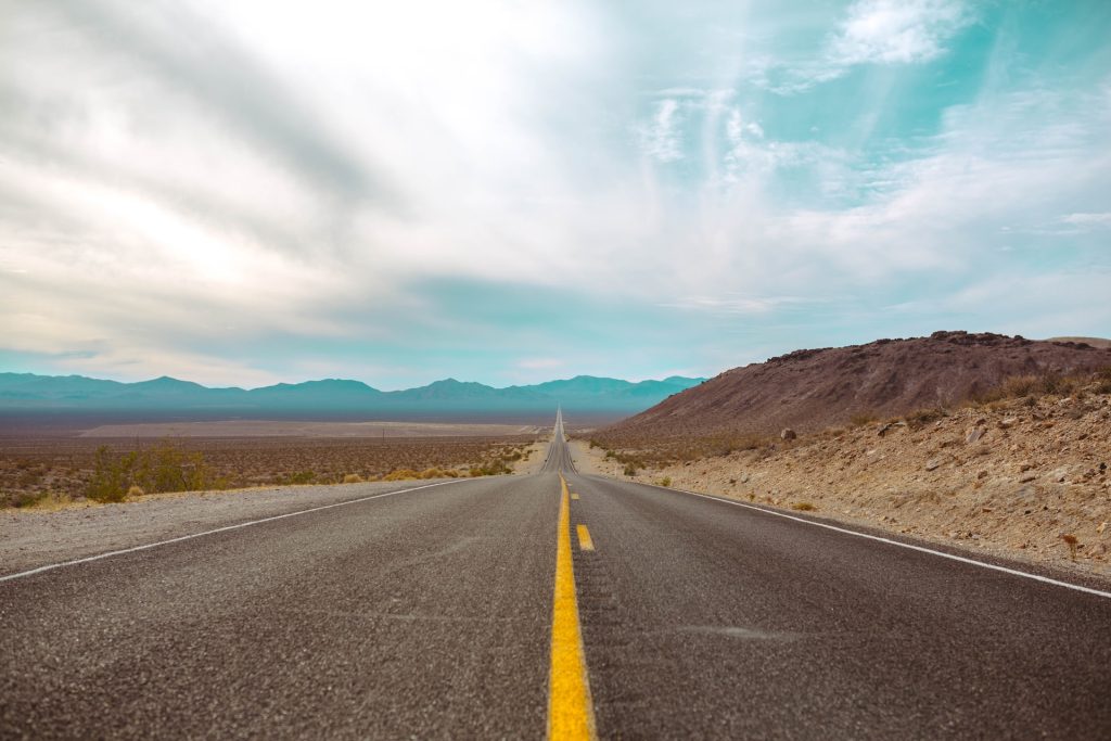 A straight road in a desert landscape stretches away from us, rising and falling over the ground.