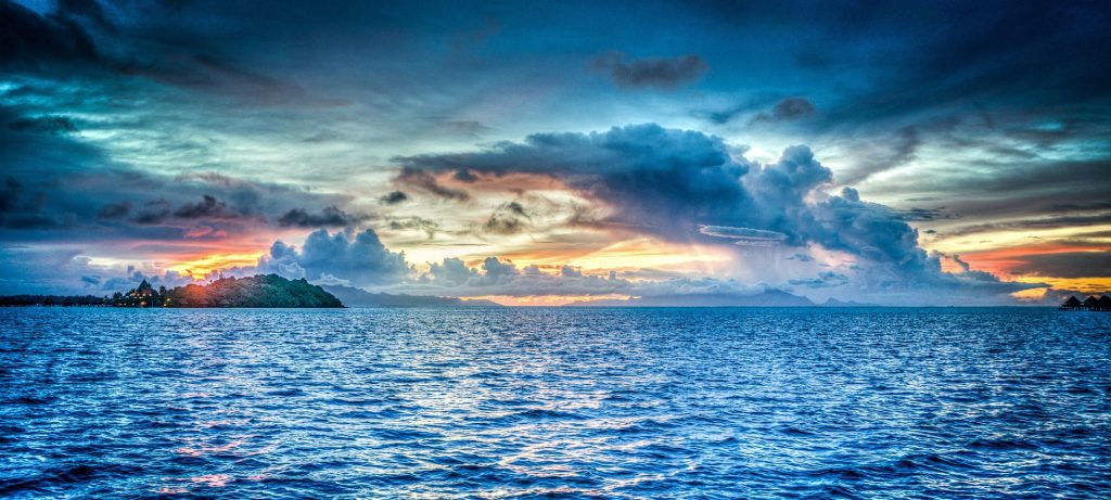 Blue sea underneath a cloudy blue sky, sunrise or sunset in the distance.