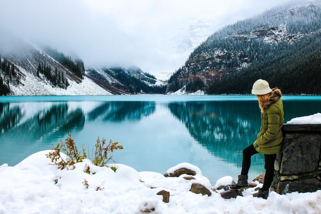 A hiker standing by a calm lake on a snowy day, with mountains in the background.