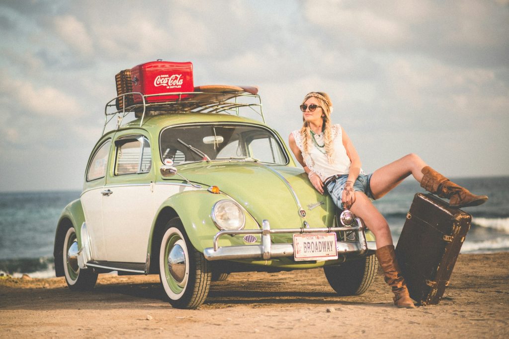 A woman reclined on the bonnet of a VW Beetle, on a beach on a sunny day.
