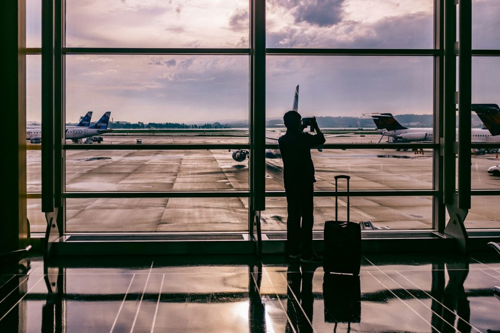 A man standing in a departure lounge, silhouetted against large windows, recording the planes on his phone.
