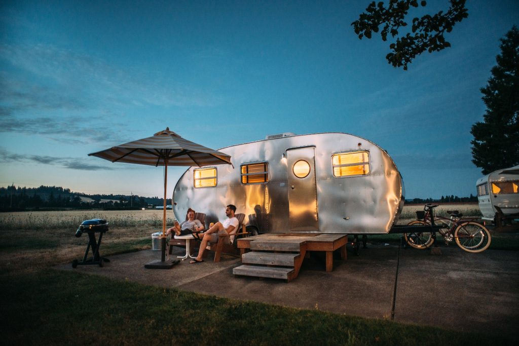 A couple relaxes under an umbrella beside a trailer at dusk, with a grill and bikes nearby.