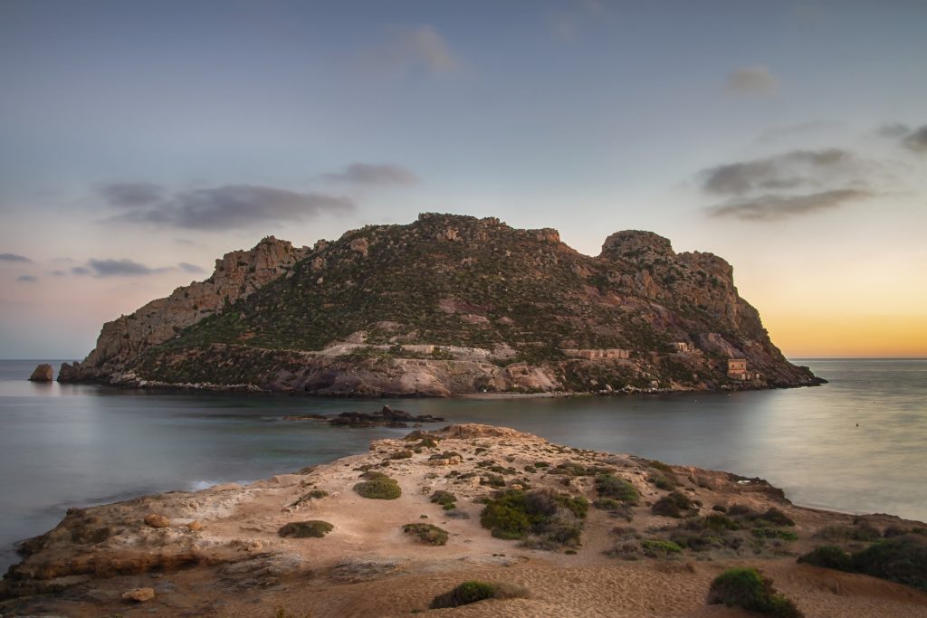 A rocky island surrounded by dark blue water, set against a clear sky.