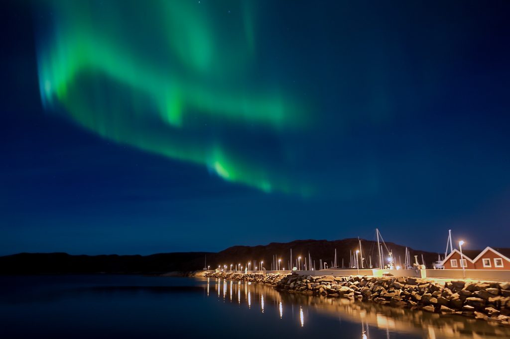 Vibrant northern lights above a calm harbor with illuminated houses and street lights along the waterfront at night.