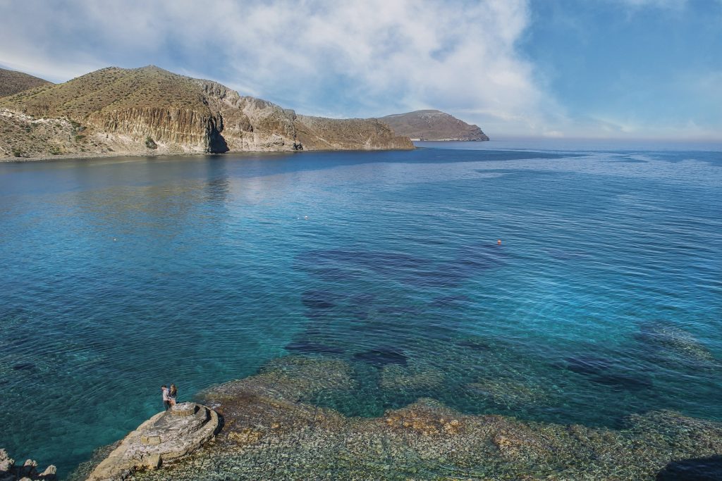 Crystal-clear turquoise waters with rocky cliffs and a lone figure standing on a stone outcrop under a bright blue sky.