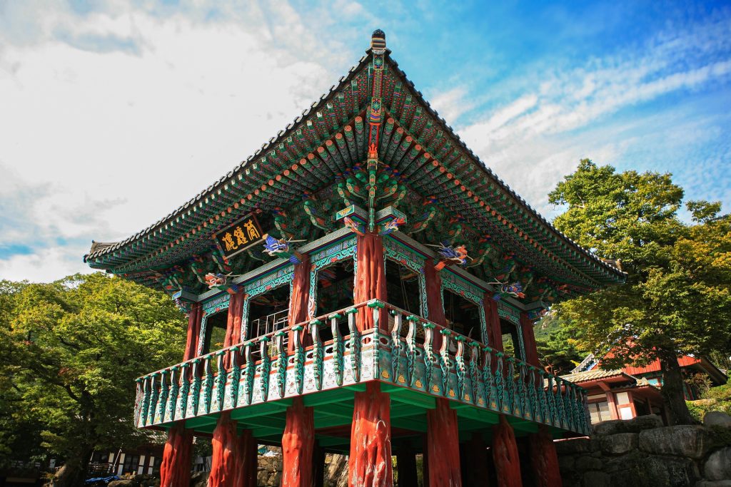 Vibrant traditional Korean pavilion with red and green painted wooden columns and a tiled roof, set against a blue sky.