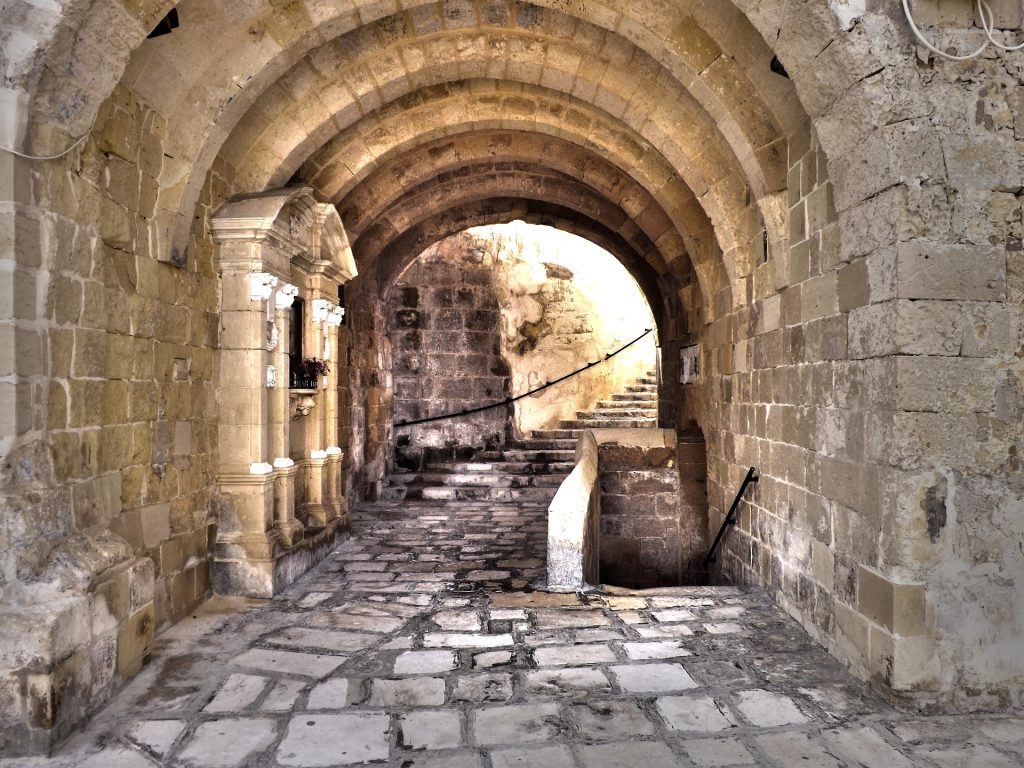 Ancient stone archway leads through a cobblestone alley, revealing historic architecture with intricate carvings.