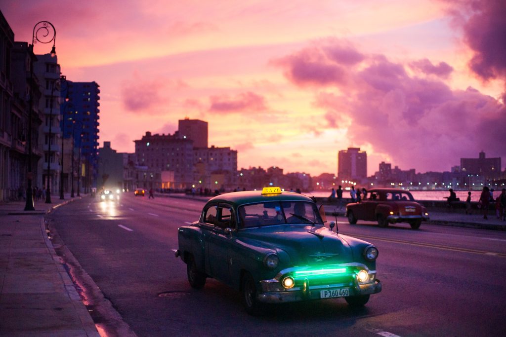 Vintage green taxi drives on a city street at sunset, with a pink and purple sky over an urban skyline.