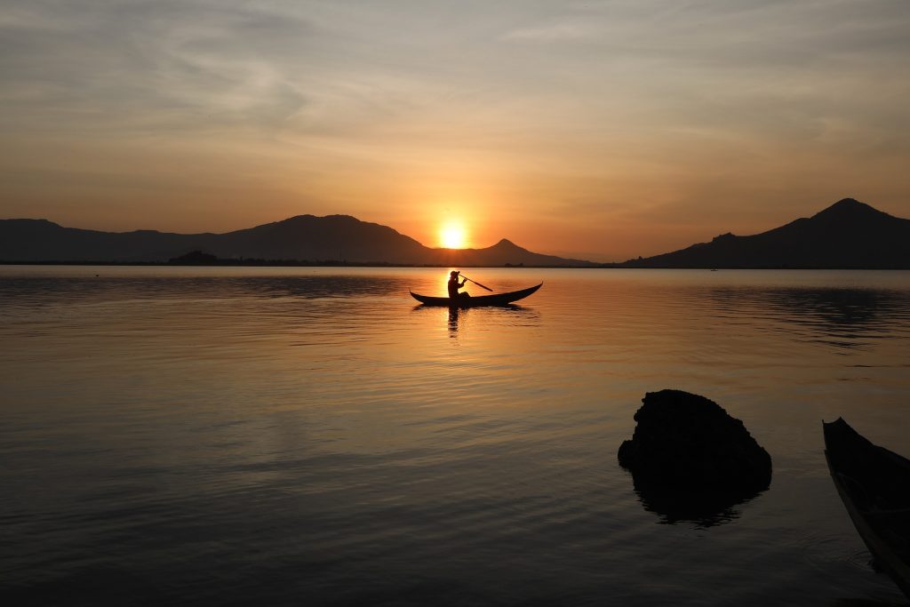 Silhouette of a person paddling a canoe on calm waters at sunset, surrounded by mountains.