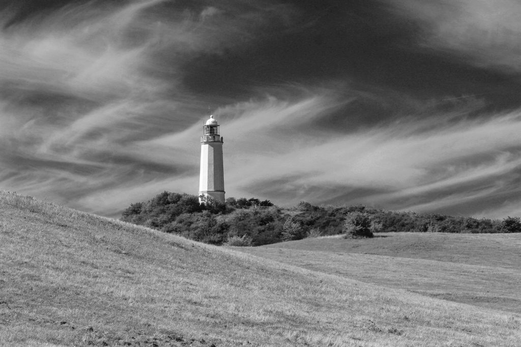 Black and white image of a solitary lighthouse atop a grassy hill, under a dramatic, swirling cloud-filled sky.
