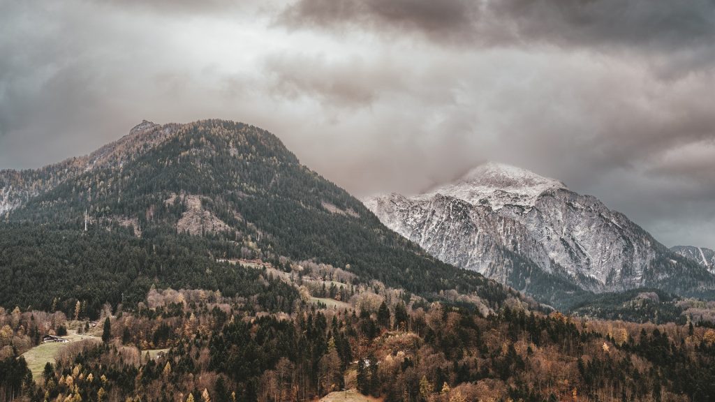 Dramatic clouds hover over snow-capped mountains and a forested valley, creating a moody and rugged landscape.