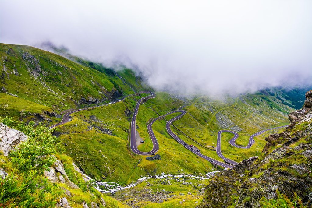 Winding mountain road weaving through lush green hills, partially covered by misty clouds.