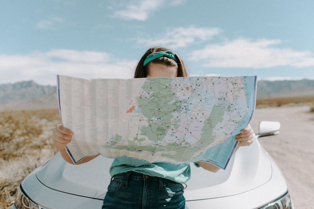 Person standing in front of a car in a desert landscape, holding a large map covering their face.