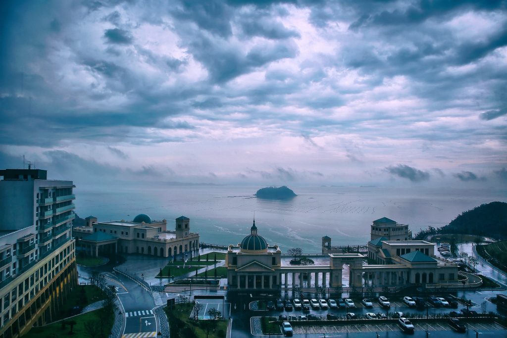 Moody view of a coastal cityscape under a cloudy sky, with buildings leading to the sea and a distant island.