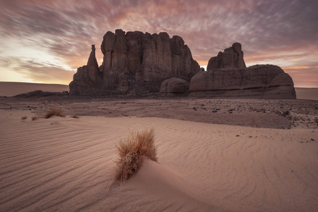 Sunrise casts a soft glow on a rugged rock formation rising majestically from a sandy desert landscape.