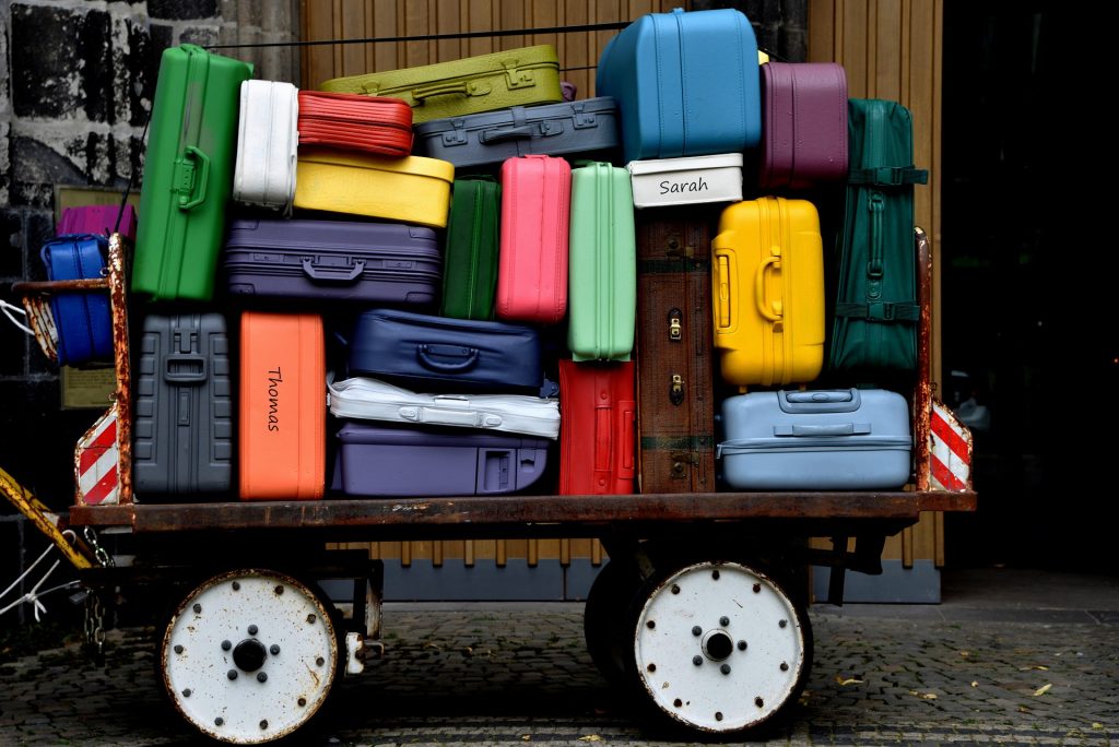 A cart overflowing with stacked, colorful suitcases in various sizes and styles, in front of a dark, textured background.