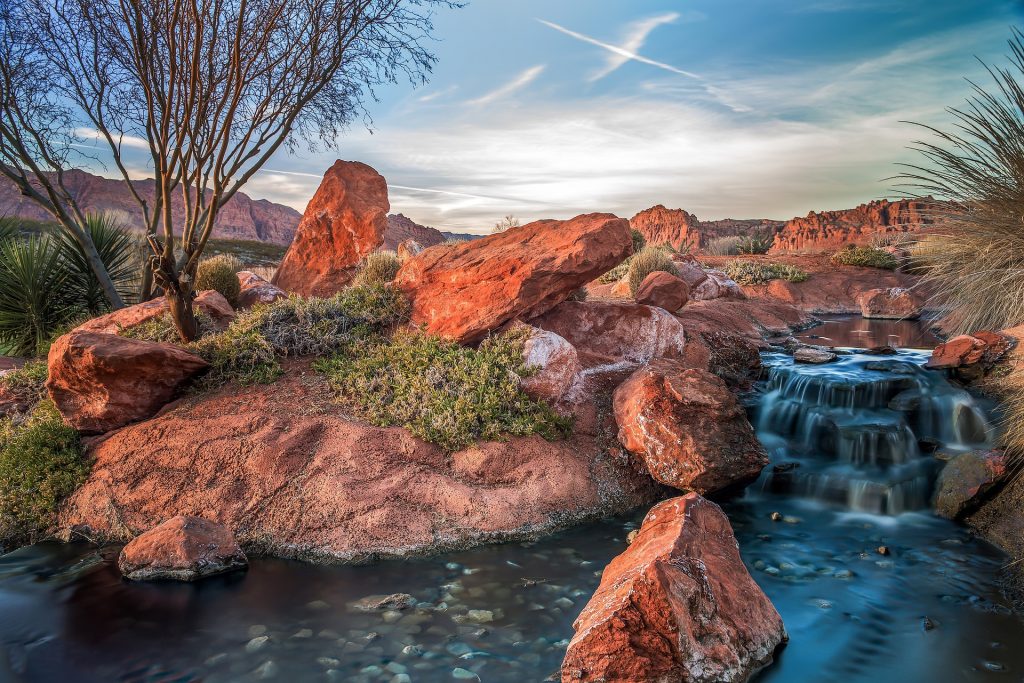 Desert oasis with red rock formations, a small waterfall flowing into a clear pool, surrounded by desert plants.