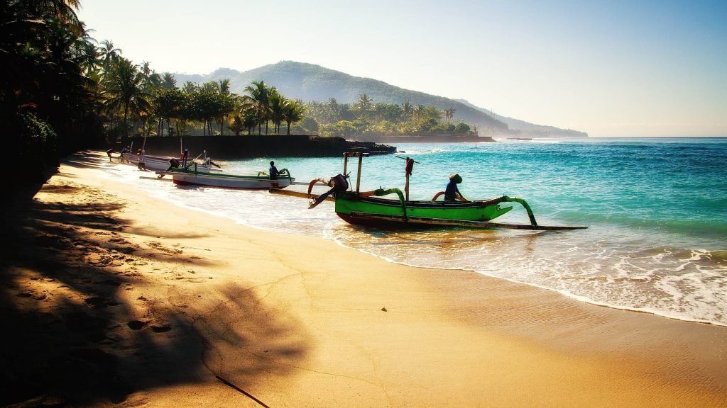Sunny beach with golden sand and turquoise waters, traditional fishing boats on the shore, framed by palm trees.