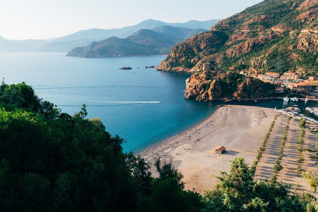Scenic coastal view of a serene bay surrounded by mountains, clear blue water, a sandy beach and a small dock with boats.
