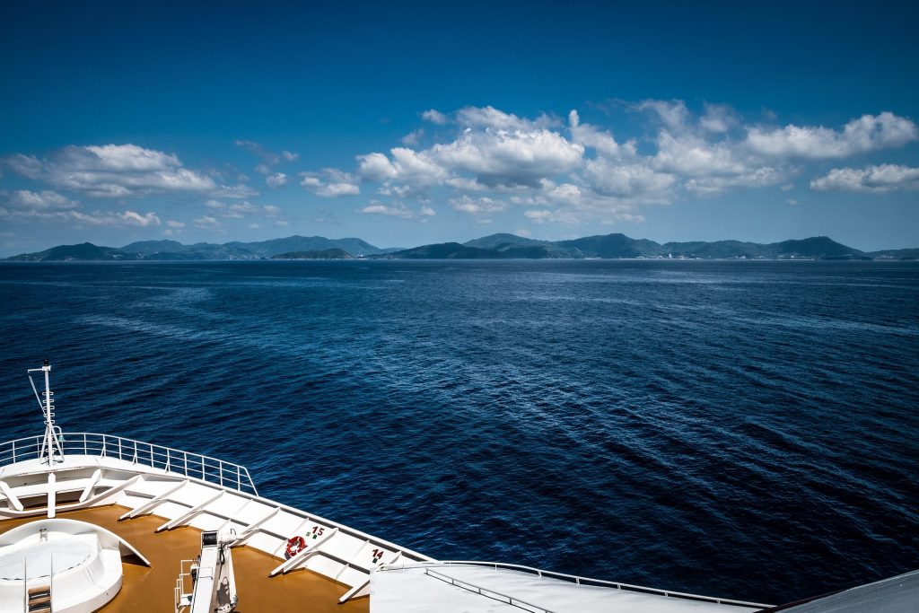 View from a ship's deck overlooking vast blue ocean waters with distant mountains under a clear, partly cloudy sky.