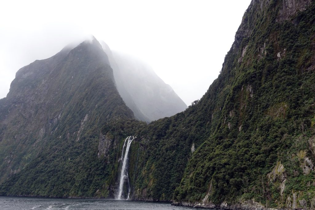Fog-covered mountain landscape with a tall, narrow waterfall cascading down lush, green cliffs into a calm body of water.