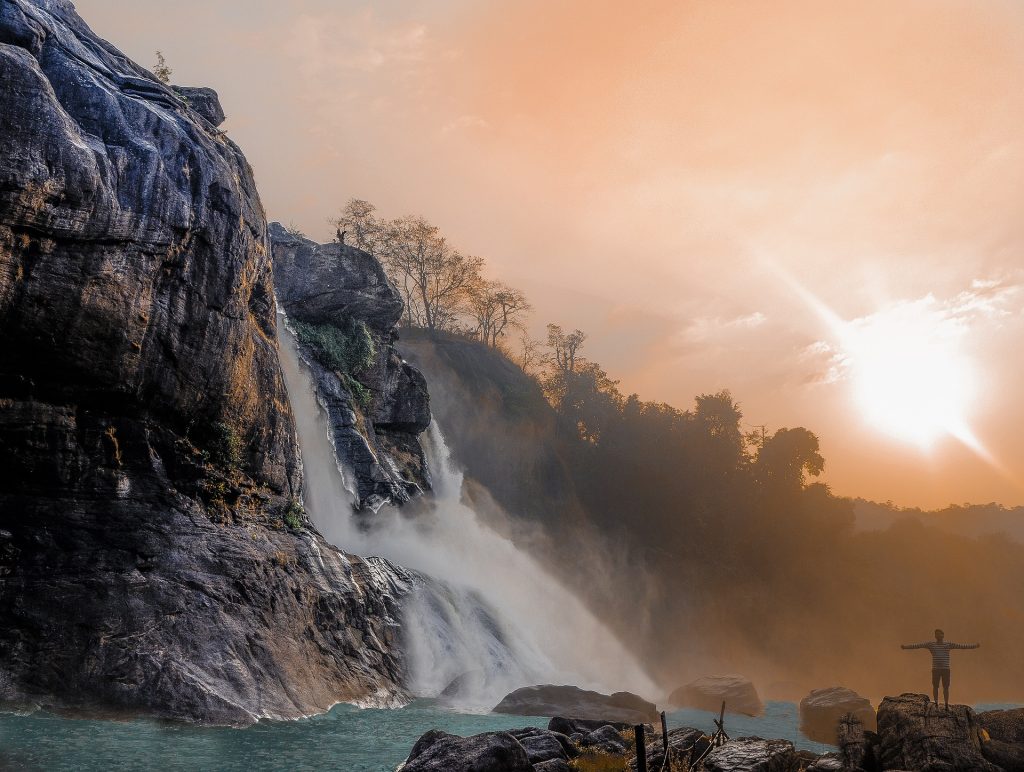 Majestic waterfall cascading down rocky cliffs into a serene pool, with a person standing in awe at sunrise.
