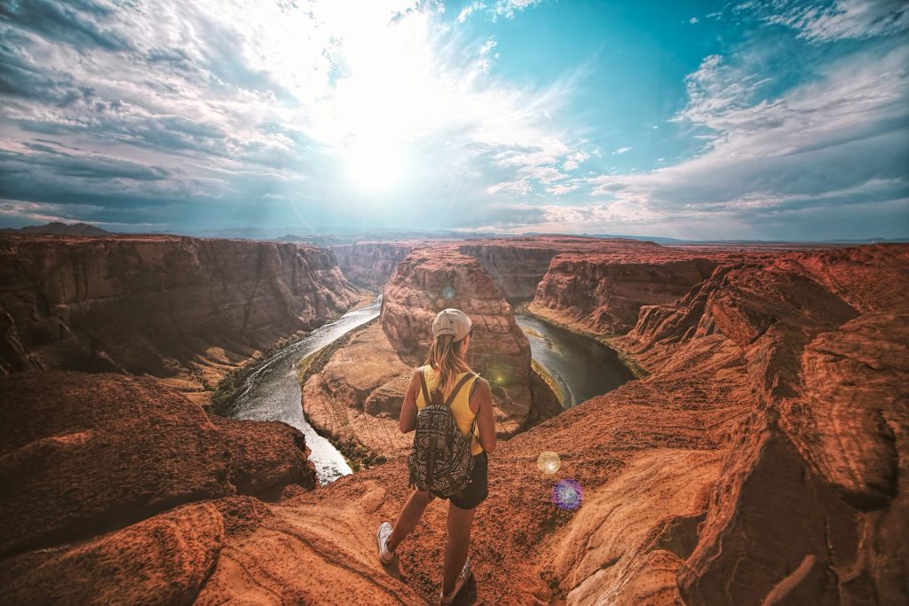 A person in a yellow tank top and backpack stands on a rocky canyon edge, gazing at a river under sunlight.