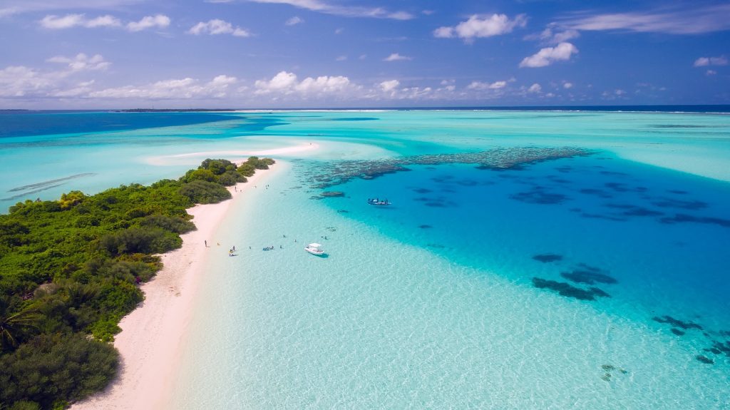 Aerial view of a tropical beach with white sand, turquoise water, lush vegetation, boats, and people walking along the shore.