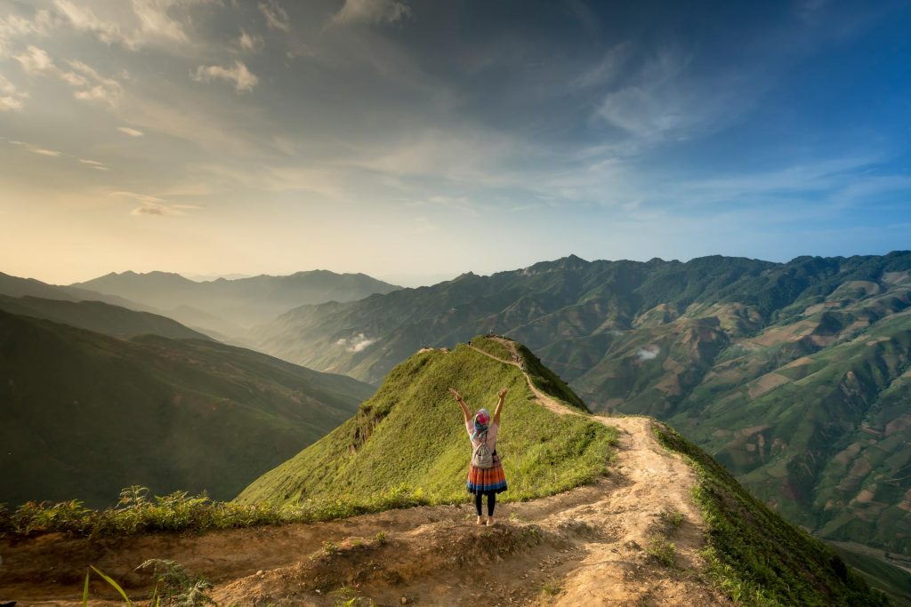 A person stands triumphantly on a mountain ridge, with lush hills and a glowing sunset in the background.