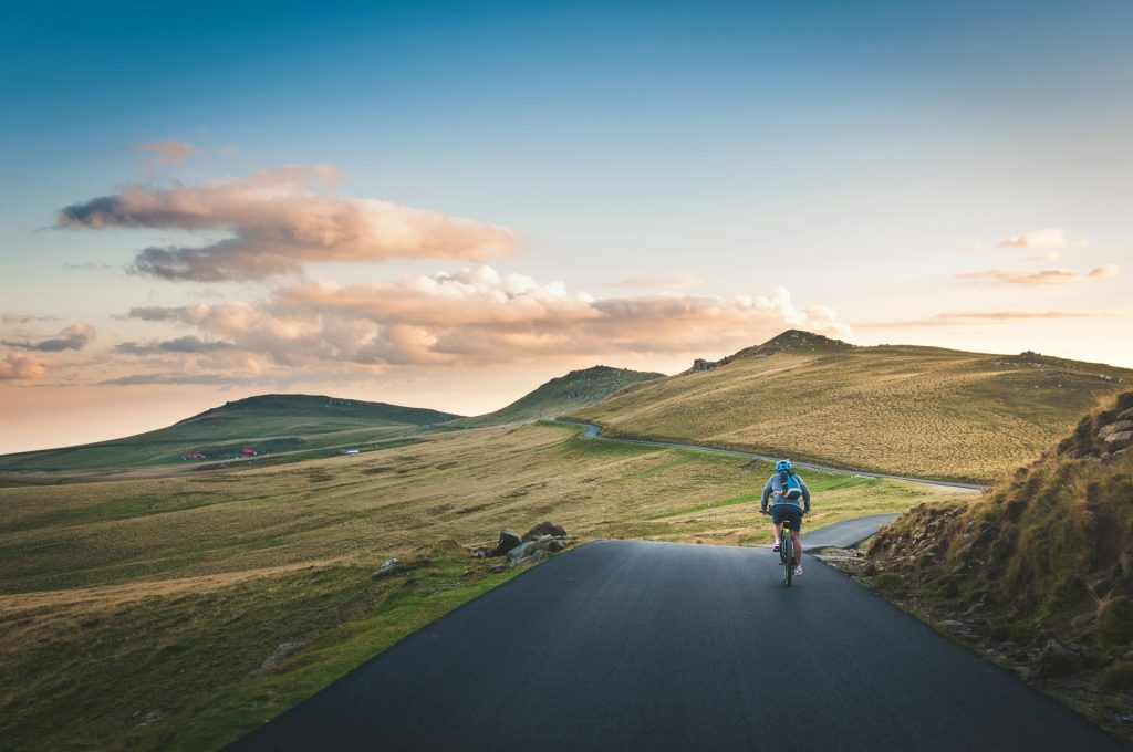Cyclist riding on a winding road through grassy hills, bathed in soft evening light with distant red structures.