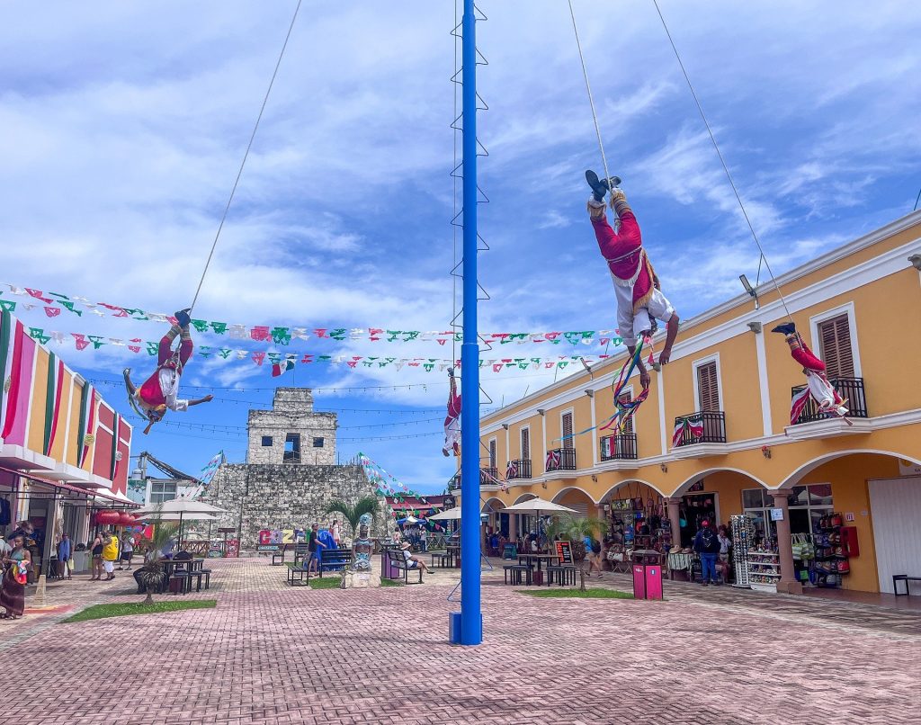 Traditional Mexican aerial performance with dancers suspended on ropes, set in a colorful plaza with historic buildings.