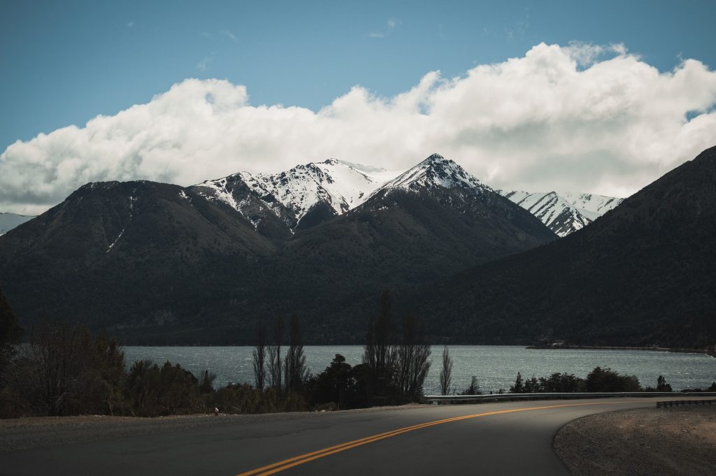 Snow-capped mountains overlooking a serene lake with a winding road in the foreground under a partly cloudy sky.