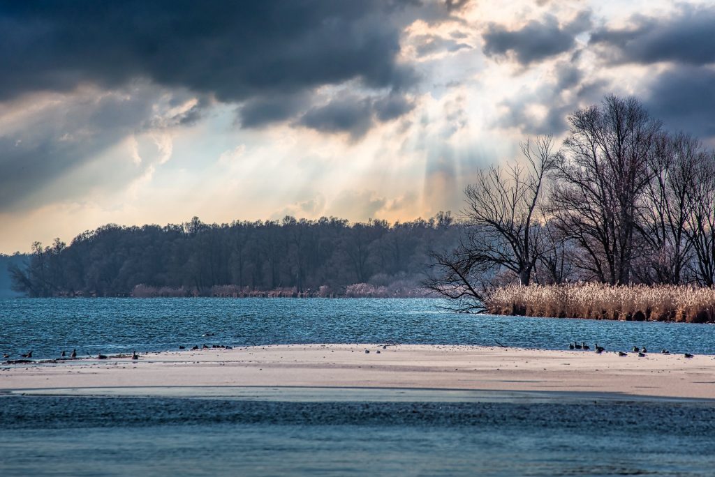 Serene lakeside scene with sun rays breaking through clouds, illuminating water, trees, and a sandy shore.