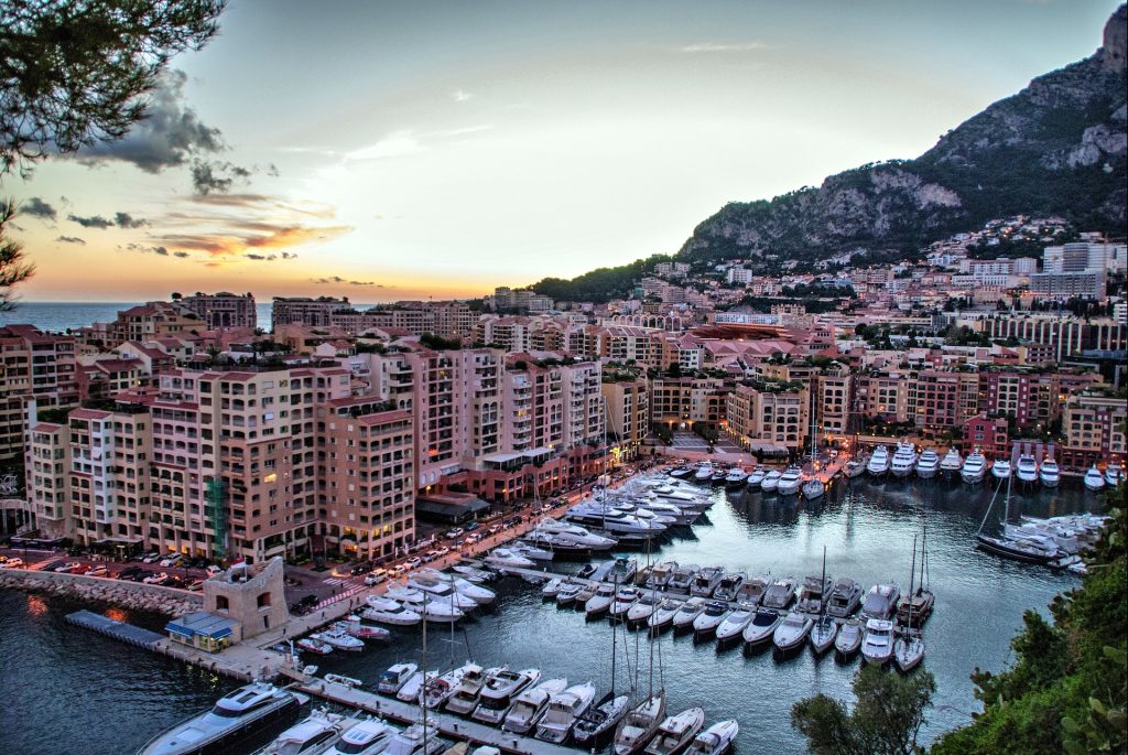 Scenic view of Monaco's marina at sunset, featuring luxury yachts, waterfront buildings, and surrounding hills.