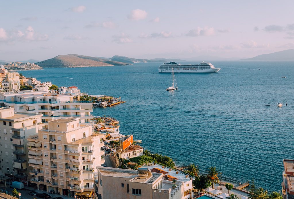 A coastal city with white buildings, a calm sea, a cruise ship, a sailboat, and distant green hills.