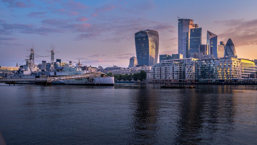 Skyline of London at dusk featuring modern skyscrapers and the historic HMS Belfast on the River Thames.