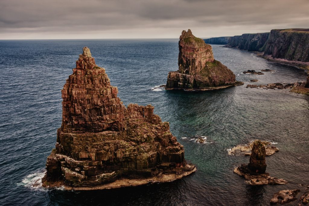 Rocky sea stacks rising from the ocean along a dramatic coastal landscape under an overcast sky.