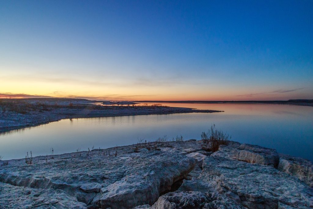 A serene lake at sunset with a clear sky, rocky foreground, and calm water reflecting the fading light.