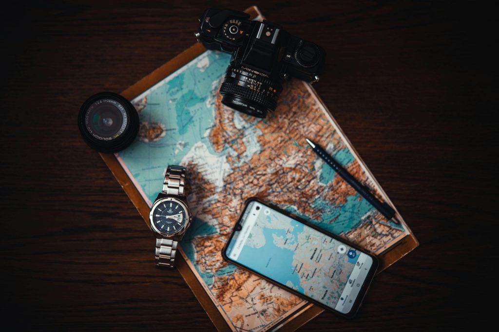 A map with a camera, watch, smartphone displaying a map, pen, and a lens—travel essentials on a wooden surface.