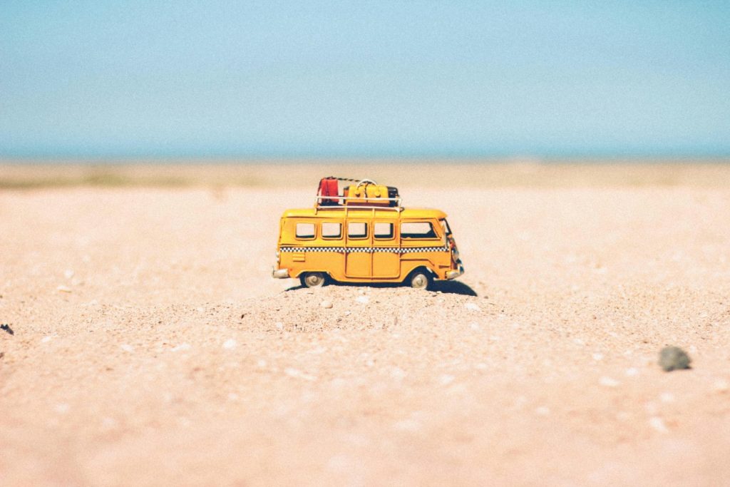 A miniature yellow bus with luggage on top sits on a sandy surface, symbolizing travel and adventure under a clear sky.