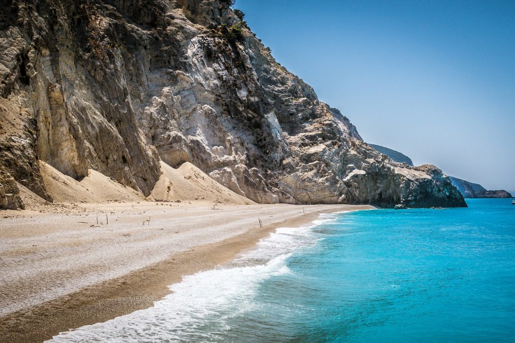 Tranquil beach with yellow sand, gentle waves, rocky cliffs, and a clear blue sky. Peaceful atmosphere.