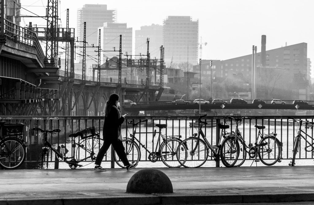 A person walks along a riverfront beside parked bicycles, with a city skyline in the background, captured in black and white.