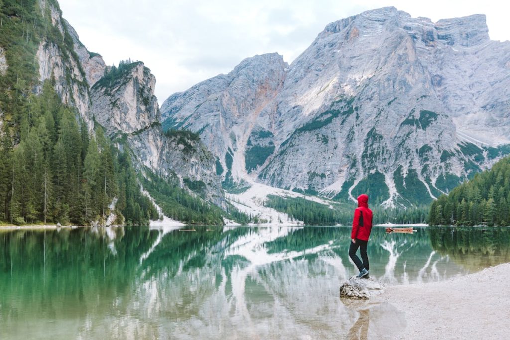 Person in a red hoodie stands on a rock by a serene lake, reflecting mountains and pine forests. Peaceful and adventurous.