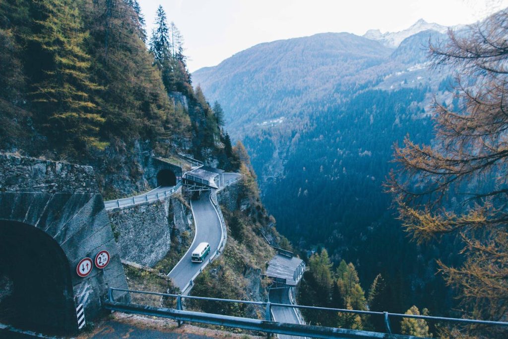A white van drives along a winding mountain road surrounded by cliffs and pine forests, leading to two tunnels.